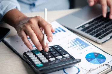 Finance analyst using a calculator while working on a budget report with charts on a laptop in the office, reviewing data, accounting, numbers, analysis, planning, costs, and profit for decision-making.