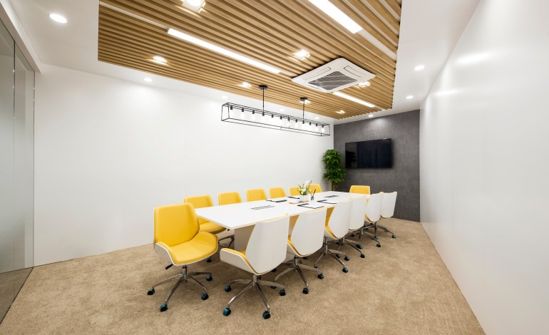 Meeting room equipped with a large table, yellow office chairs, which completely surround the table, and a display mounted on one of the walls. The ceiling has an AC unit and wood panels and floors are carpeted. 