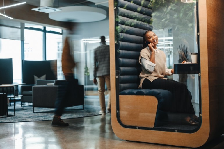 Happy businesswoman sitting in a coworking space, speaking on her smartphone, discussing a business call. Smiling and successful, showcasing her communication and remote working skills.