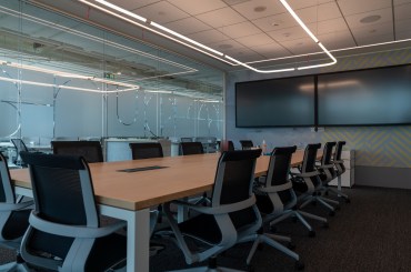 Business meeting room with a large desk, surrounded by chairs. On one of the walls there are two black screens that are blank.