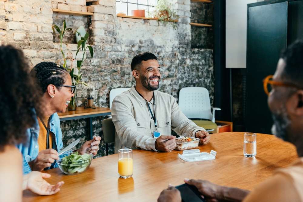 A group of colleagues shares a joyful lunch hour in a bright office with thriving indoor plants, fostering teamwork and connection over a meal, laughter, and casual conversation.