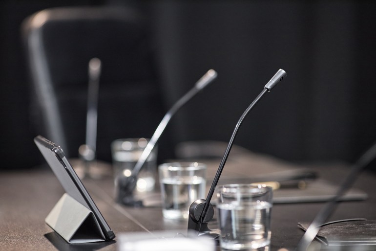 A bunch of microphones sitting on a table. Next to each microphone there is a glass of water, and in front of the microphones there is a tablet sitting upright. 