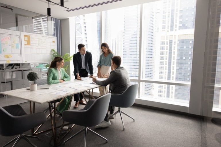 Successful company meeting space with a team of four colleagues, stakeholders, and partners sitting at a table, working together on a project and brainstorming in the workspace.