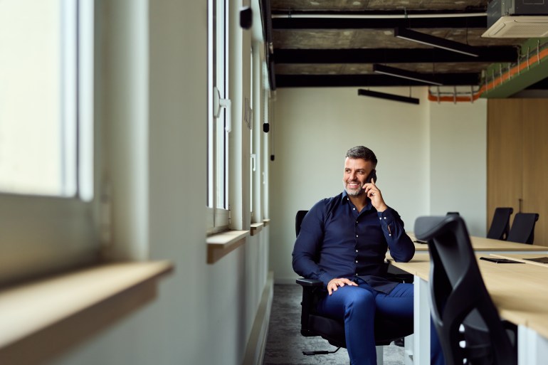 A man in a navy shirt sitting down and talking on the phone in a meeting room.