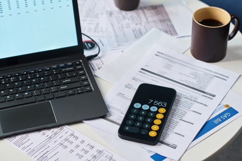 Closing the smartphone with a calculator app, alongside a laptop, a coffee mug, and assorted financial documents, including a gas bill on the desk, illustrating the process of paying taxes and managing expenses.
