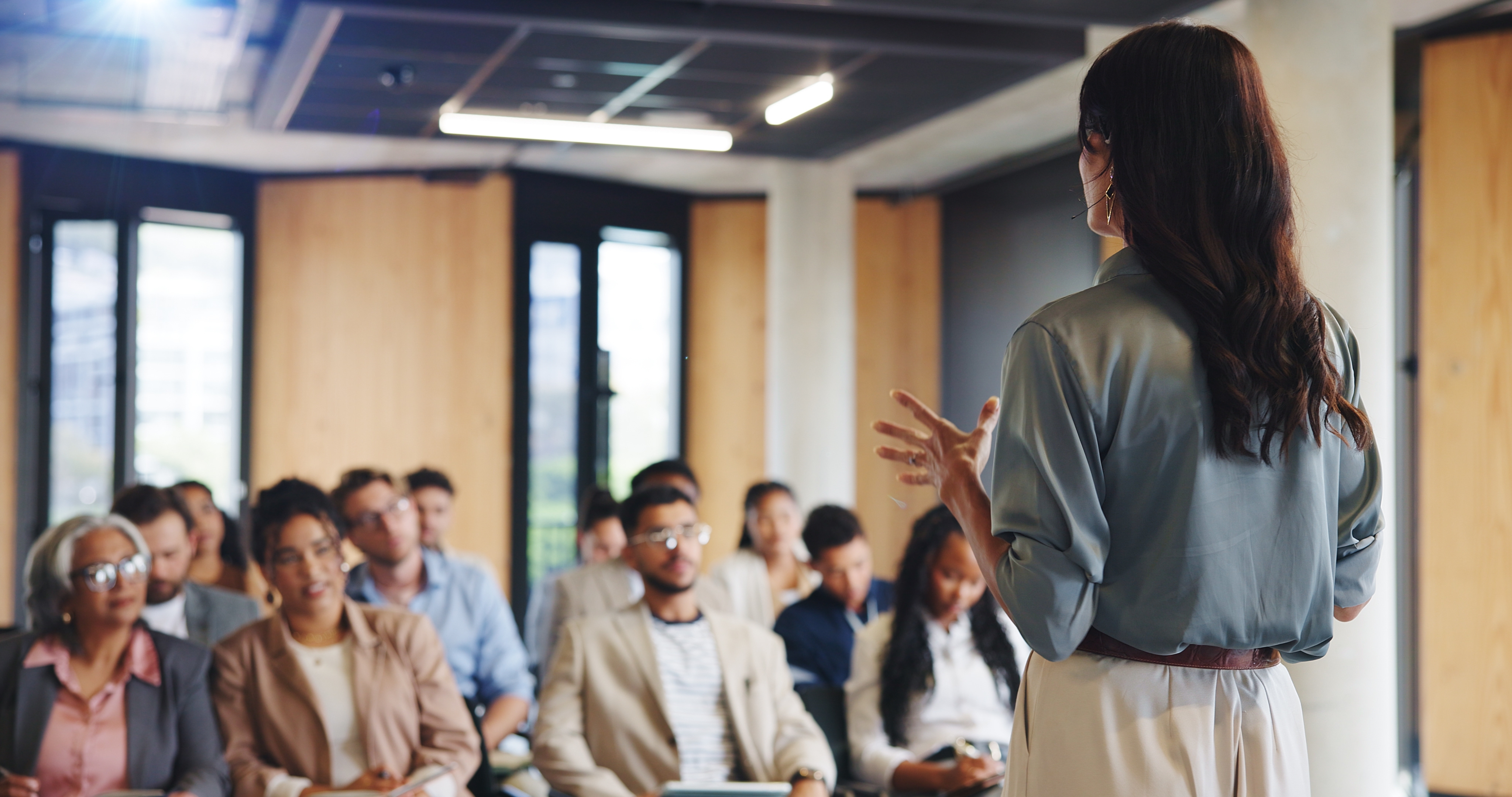 How to Run a Workshop in a Coworking Space: Room Layouts, Supplies, Sound, and Costs Woman presenting in front of a small, seated group of people, They are in a meeting room.