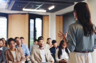 Woman presenting in front of a small, seated group of people, They are in a meeting room.