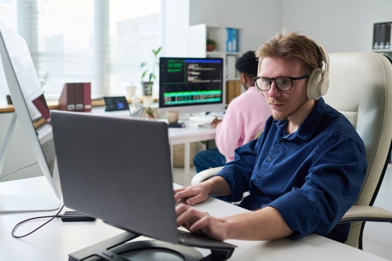 A man wearing headphones, working on a laptop in an office, sitting at a desk and focusing on programming while another person works on a computer in the background. 