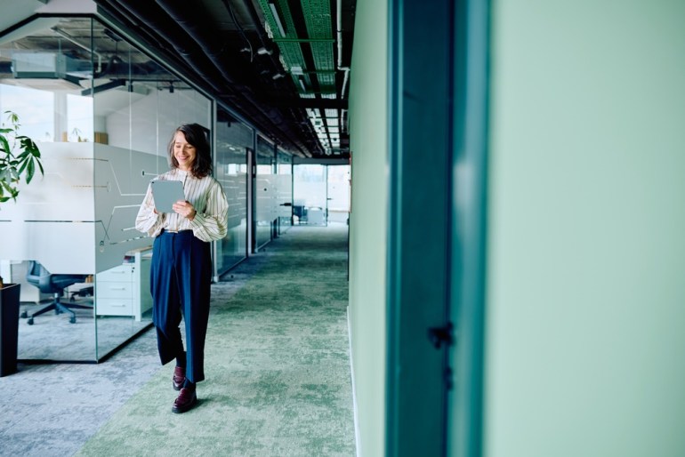 Businesswoman walking through a contemporary office corridor, smiling while using a digital tablet.