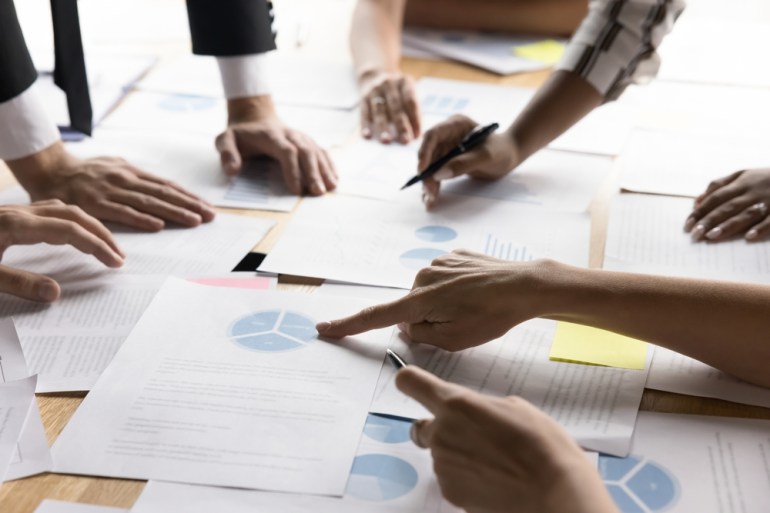 Close-up of business professionals pointing at various charts and documents spread across a conference table, participants engaged in discussions, analysis, reviewing graphs and pie charts, focused on data analysis.
