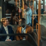 An image focusing on two people sitting in the bus, talking. They are smiling and surrounded by other people taking the bus.