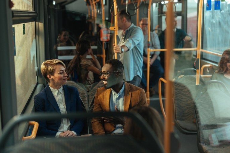 From a Drive Away to a Walk Away: Best U.S. Cities to Live in Without a Car An image focusing on two people sitting in the bus, talking. They are smiling and surrounded by other people taking the bus.